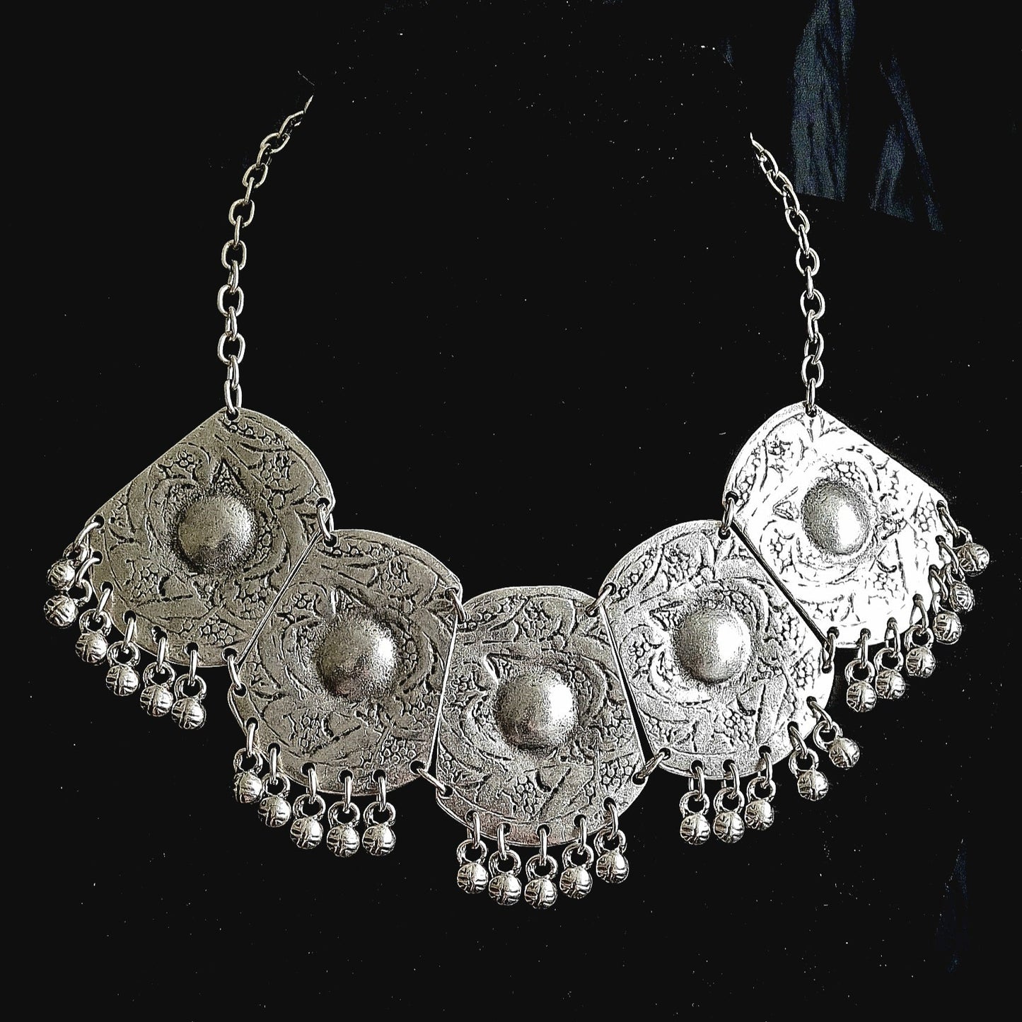 Intricate artisan-crafted silver bohemian bib necklace featuring five engraved circular medallions with hanging metallic charms, isolated on a deep black background.