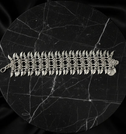 Luxury product display of a wide silver spike cuff bracelet laid flat on a circular black marble plinth, showcasing the intricate metalwork and bohemian-inspired charms.