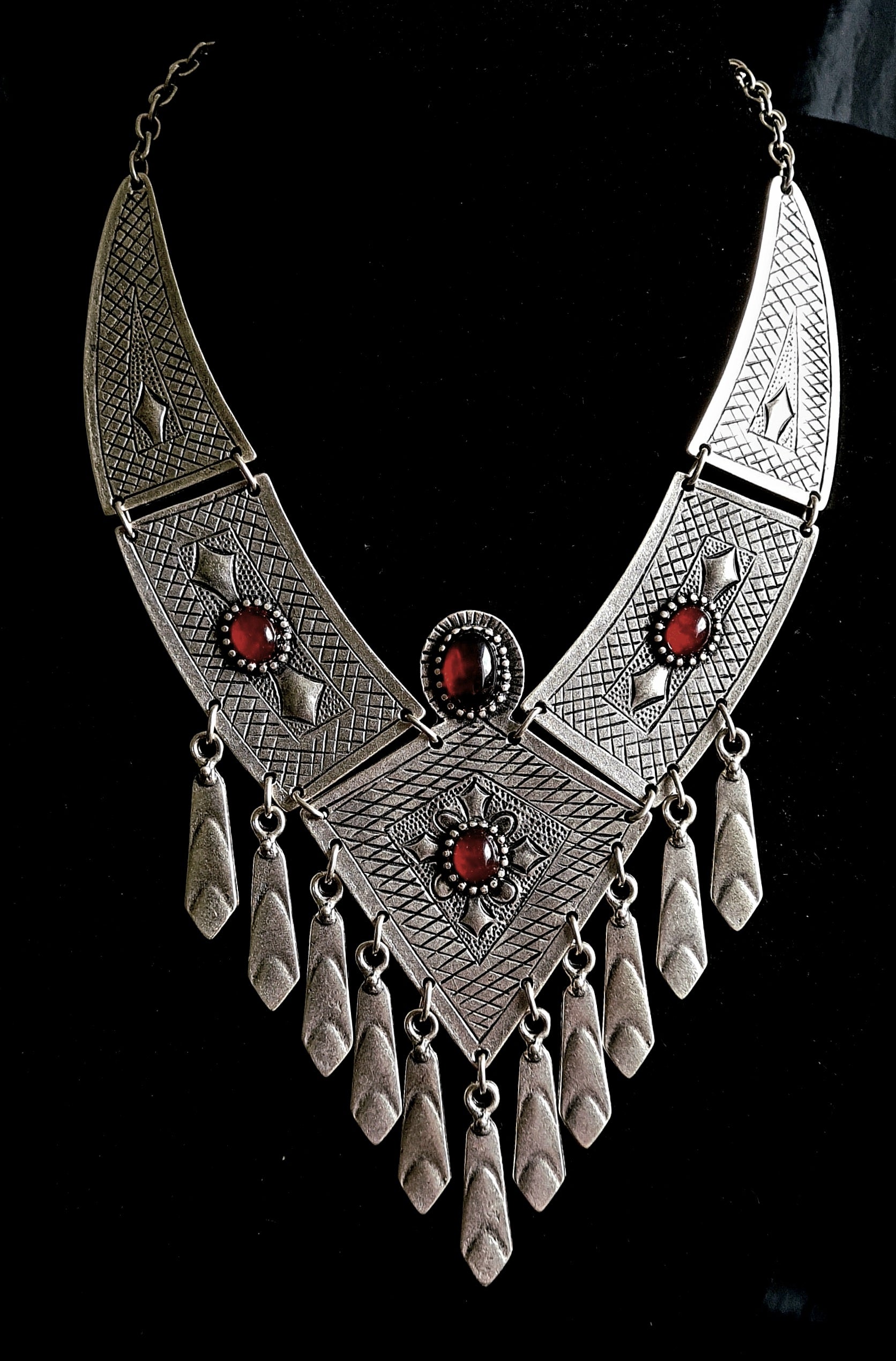 Intricate artisan-crafted sterling silver tribal bib necklace with deep red gemstone accents and hanging charms, isolated on a seamless white background for a premium catalog display.