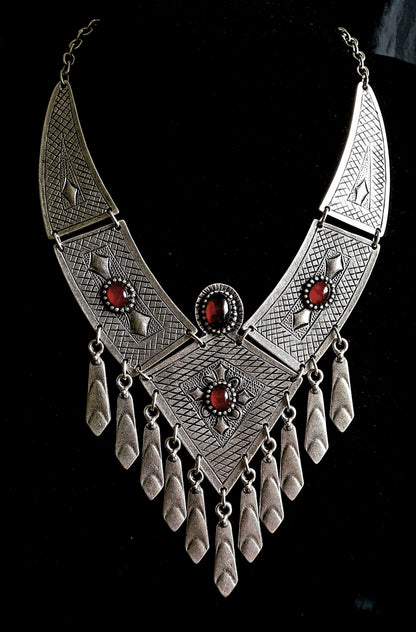 Intricate artisan-crafted sterling silver tribal bib necklace with deep red gemstone accents and hanging charms, isolated on a seamless white background for a premium catalog display.