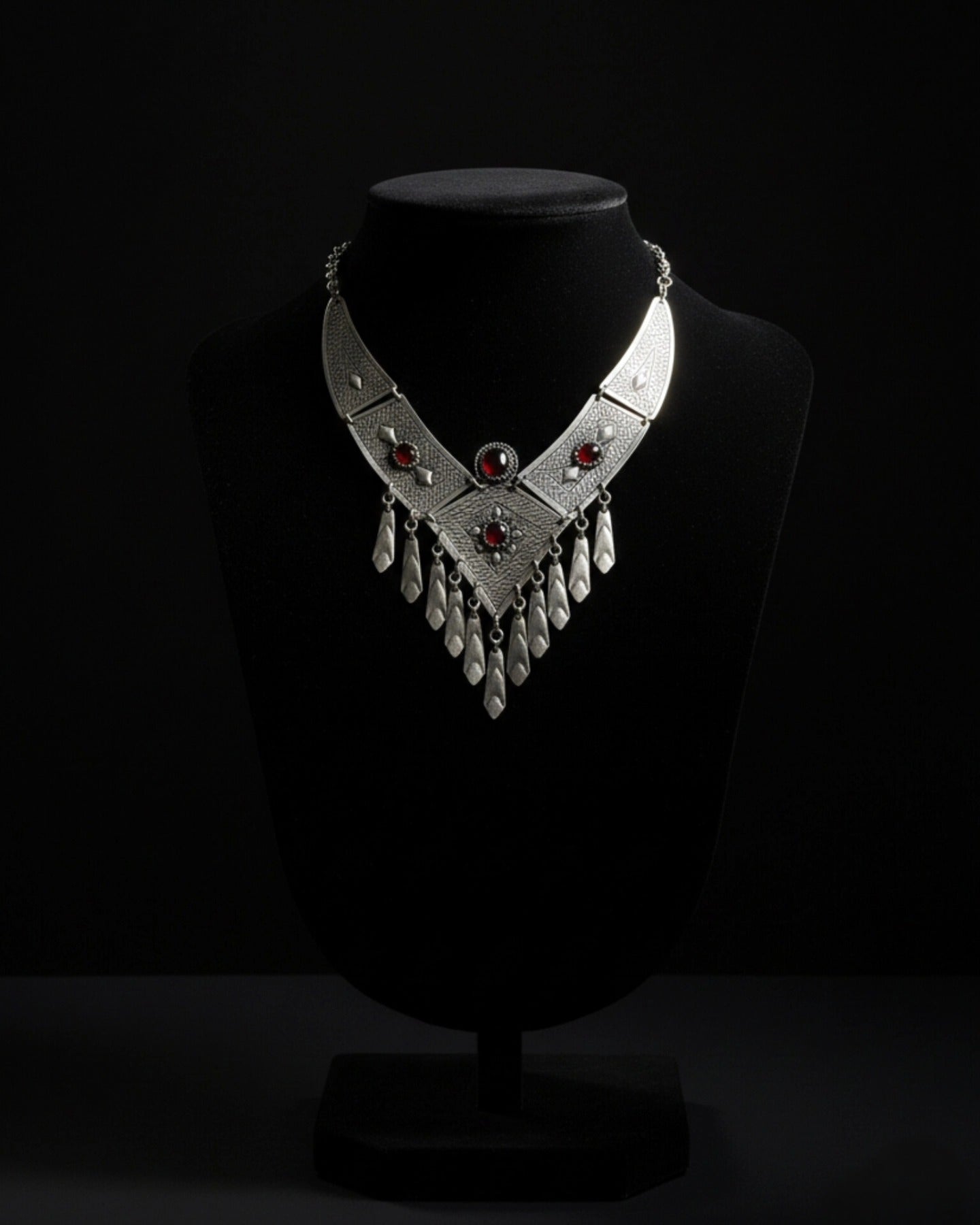 Symmetric high-fashion studio portrait of a model wearing an intricate, large silver tribal-style bib necklace accented with deep red gemstones, set against a dark, minimalist background.