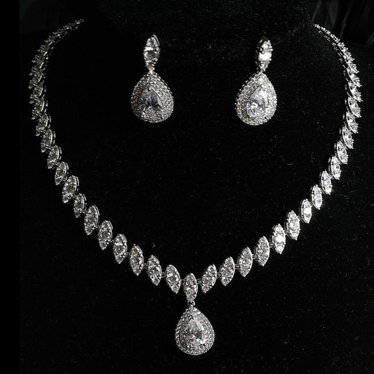 A necklace and earrings set with diamonds on a black background. The necklace is a long, silver chain with a large diamond pendant. The earrings are drop earrings with pear-shaped diamonds. The background is a black cloth.