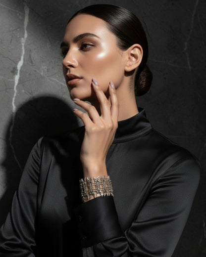 High-fashion editorial portrait of a model with a sleek bun wearing an ornate silver spike cuff bracelet and a black silk high-neck top, posed against a dark marble background with dramatic lighting.
