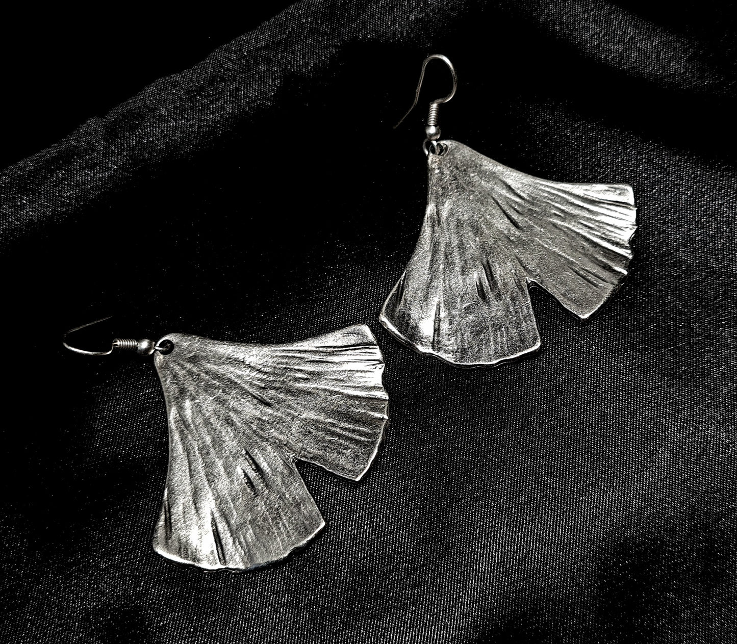 Pair of artisan-crafted silver ginkgo leaf earrings displayed on a deep black velvet background with dramatic high-fashion lighting.