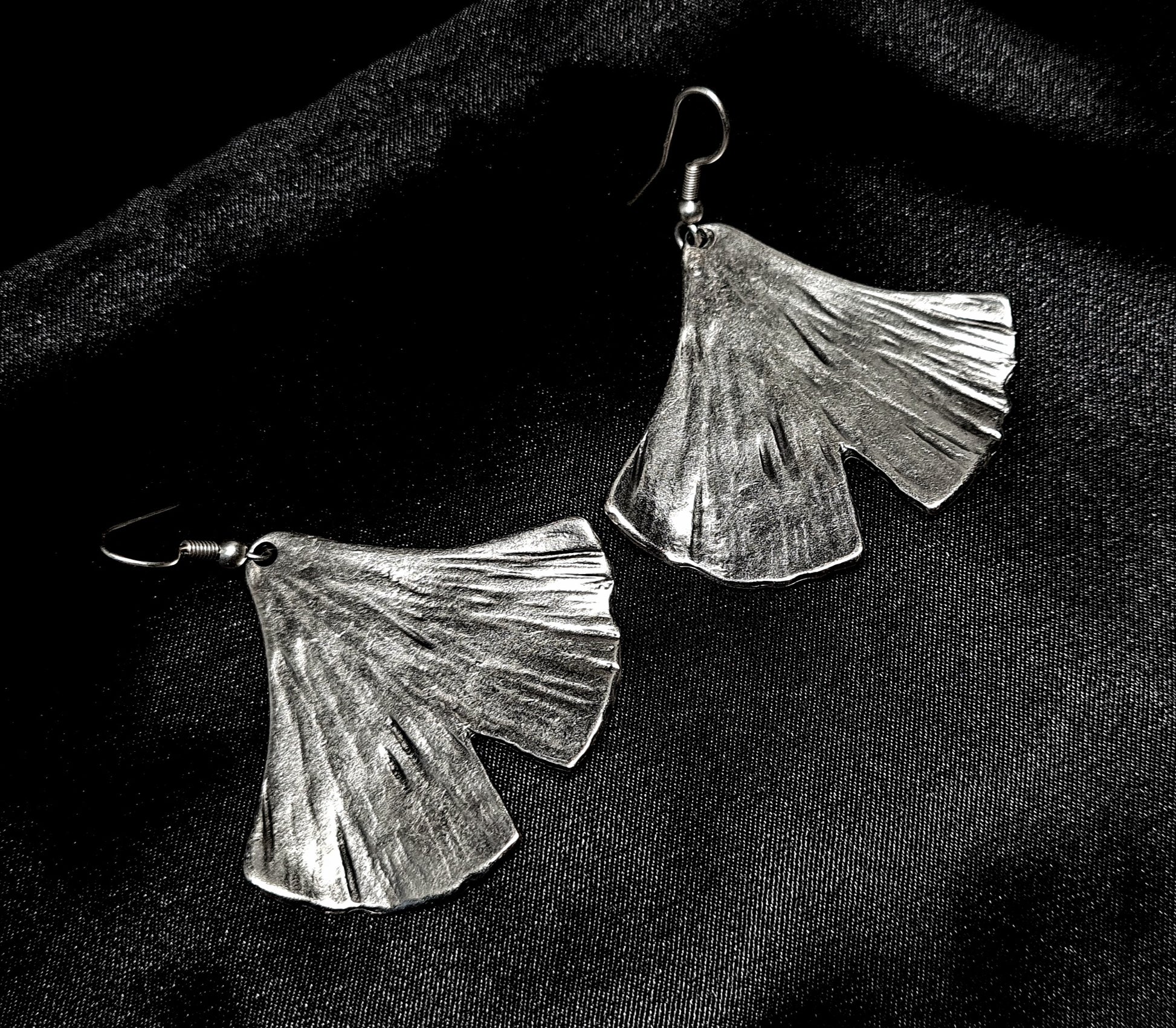 Pair of artisan-crafted silver ginkgo leaf earrings displayed on a deep black velvet background with dramatic high-fashion lighting.