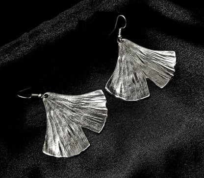Pair of artisan-crafted silver ginkgo leaf earrings displayed on a deep black velvet background with dramatic high-fashion lighting.