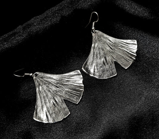 Pair of artisan-crafted silver ginkgo leaf earrings displayed on a deep black velvet background with dramatic high-fashion lighting.