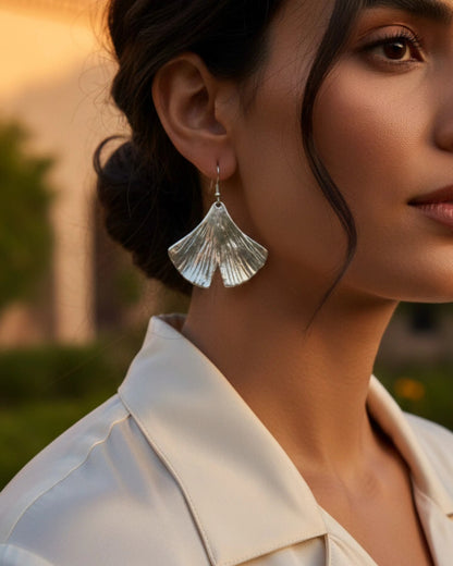 Close-up high-fashion editorial portrait of a woman wearing silver ginkgo leaf statement earrings, styled with a silk cream blouse and an elegant updo, captured in warm sunset lighting.