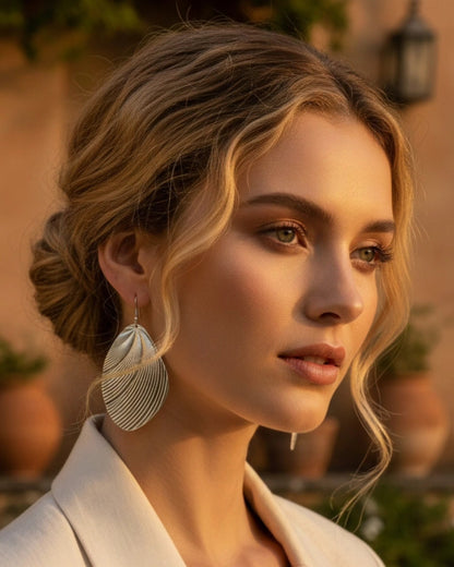An aspirational luxury campaign shot featuring a model with an elegant blonde bun and soft, face-framing waves. She is wearing oversized, sculptural silver drop earrings with a textured, pleated finish, reflecting a high-status, fashion-forward brand aesthetic for La Pasión.