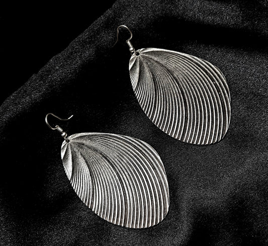 Professional product photography of a single La Pasión sculptural silver earrings on a deep black background. The image highlights the intricate pleated texture and the high-polish finish of the premium sterling silver-tone metal.