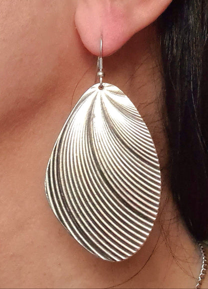 A macro, high-fashion close-up of the sculptural silver earring worn on the ear. The shot emphasizes the scale of the piece and the elegant way the pleated metal catches the light against the skin, reflecting a high-status luxury brand aesthetic.