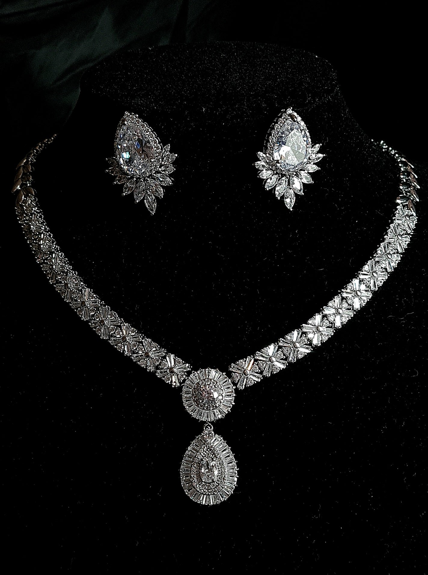 A luxury bridal jewelry set featuring a wide, intricate diamond-encrusted choker necklace with a large pear-shaped drop pendant and matching teardrop halo earrings, displayed against a premium black velvet background.
