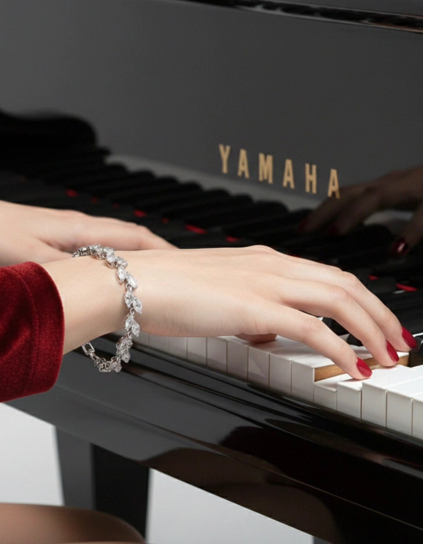 A close-up shot of hands with red nail polish playing a black Yamaha grand piano, highlighting a sparkling marquise-cut diamond tennis bracelet on the wrist.