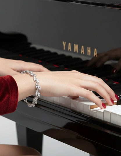 A close-up shot of hands with red nail polish playing a black Yamaha grand piano, highlighting a sparkling marquise-cut diamond tennis bracelet on the wrist.