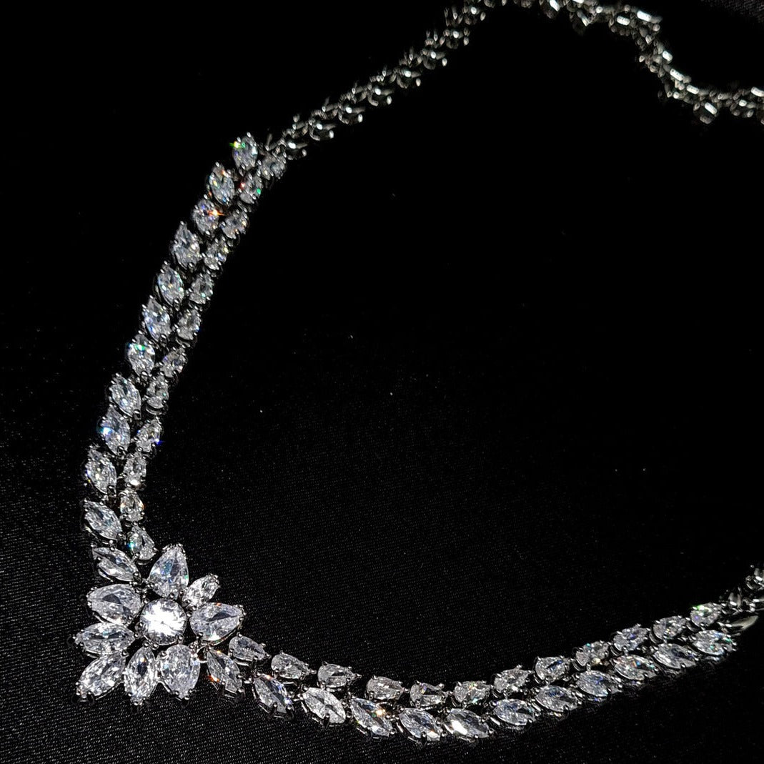 A high-detail shot of a silver-toned necklace featuring a double row of marquise-cut diamonds leading to a central floral cluster, set against a black silk background.