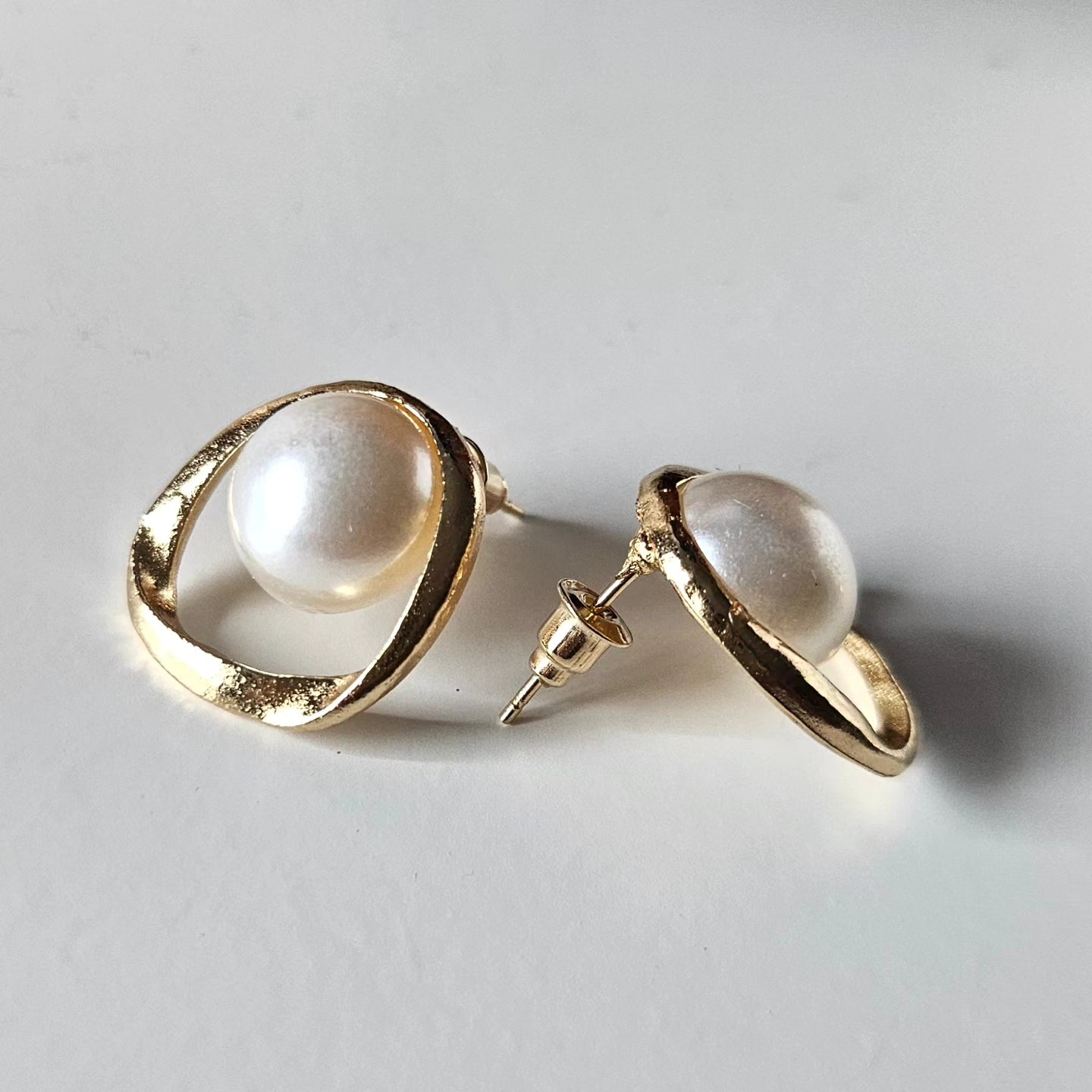 High-fashion stud earrings featuring large, lustrous white pearls encased in irregular, hand-sculpted organic gold hoops, showcasing a minimalist and sophisticated luxury aesthetic.