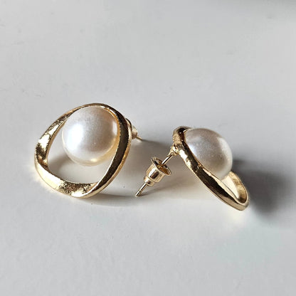 High-fashion stud earrings featuring large, lustrous white pearls encased in irregular, hand-sculpted organic gold hoops, showcasing a minimalist and sophisticated luxury aesthetic.