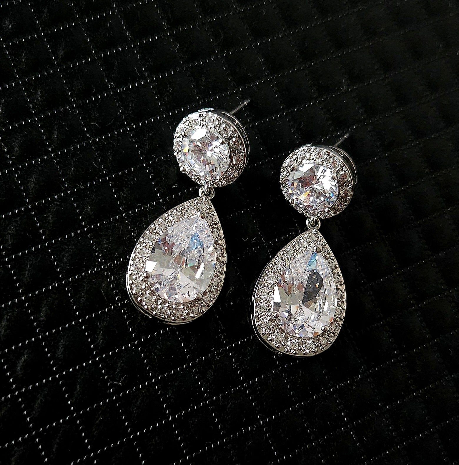 A pair of high-fashion teardrop earrings featuring a pear-cut center stone surrounded by a brilliant halo, displayed on a textured black luxury background.