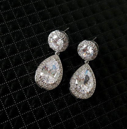 A pair of high-fashion teardrop earrings featuring a pear-cut center stone surrounded by a brilliant halo, displayed on a textured black luxury background.