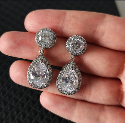 A hand-held close-up shot of elegant pear-shaped halo drop earrings, showcasing the scale and brilliant light-catching quality of the jewelry.