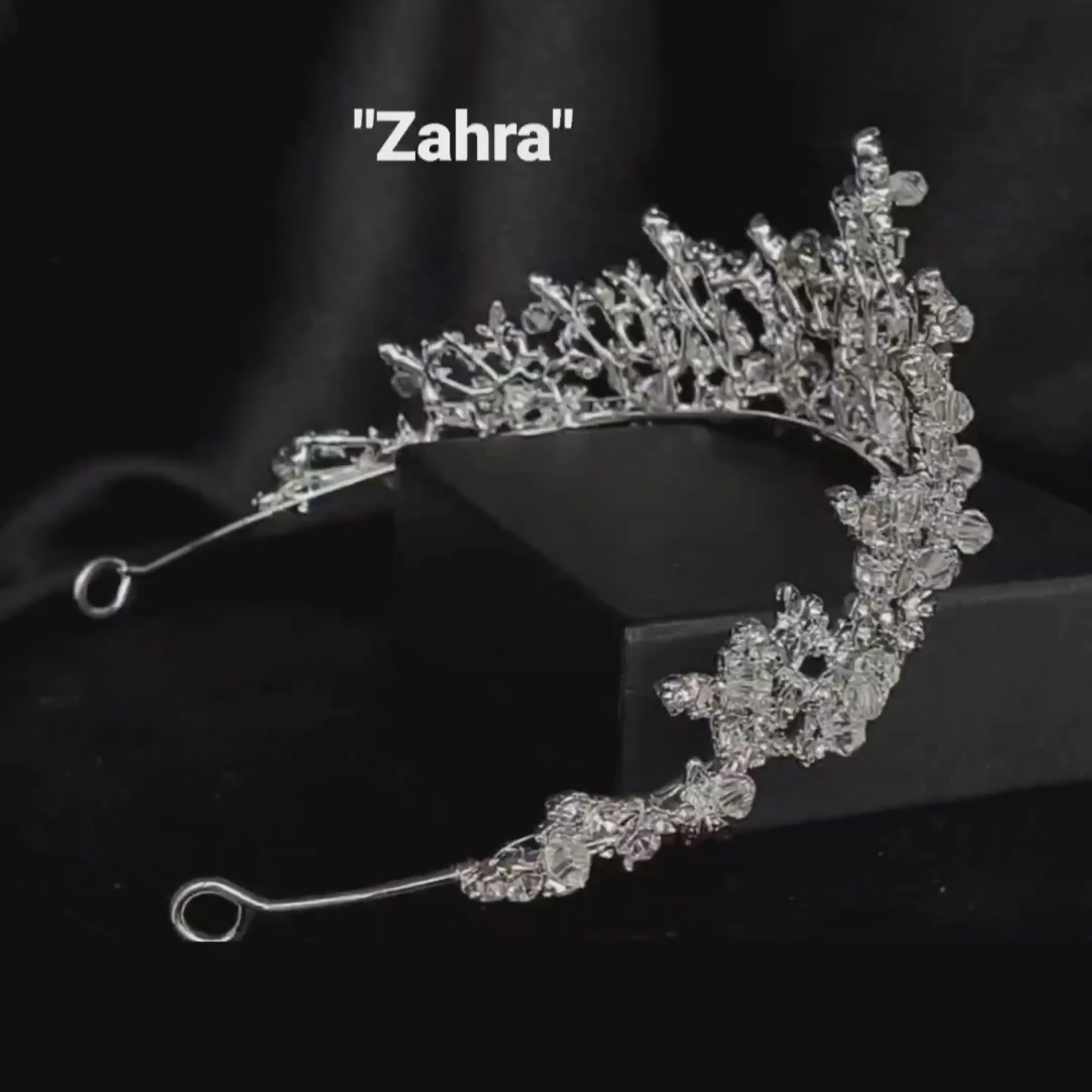 A silver tiara with rhinestones sitting on top of a black box. The tiara is made up of multiple curved bands, each of which is decorated with rhinestones. The tiara is sitting on top of a black box, which is partially obscuring the bottom of the tiara.