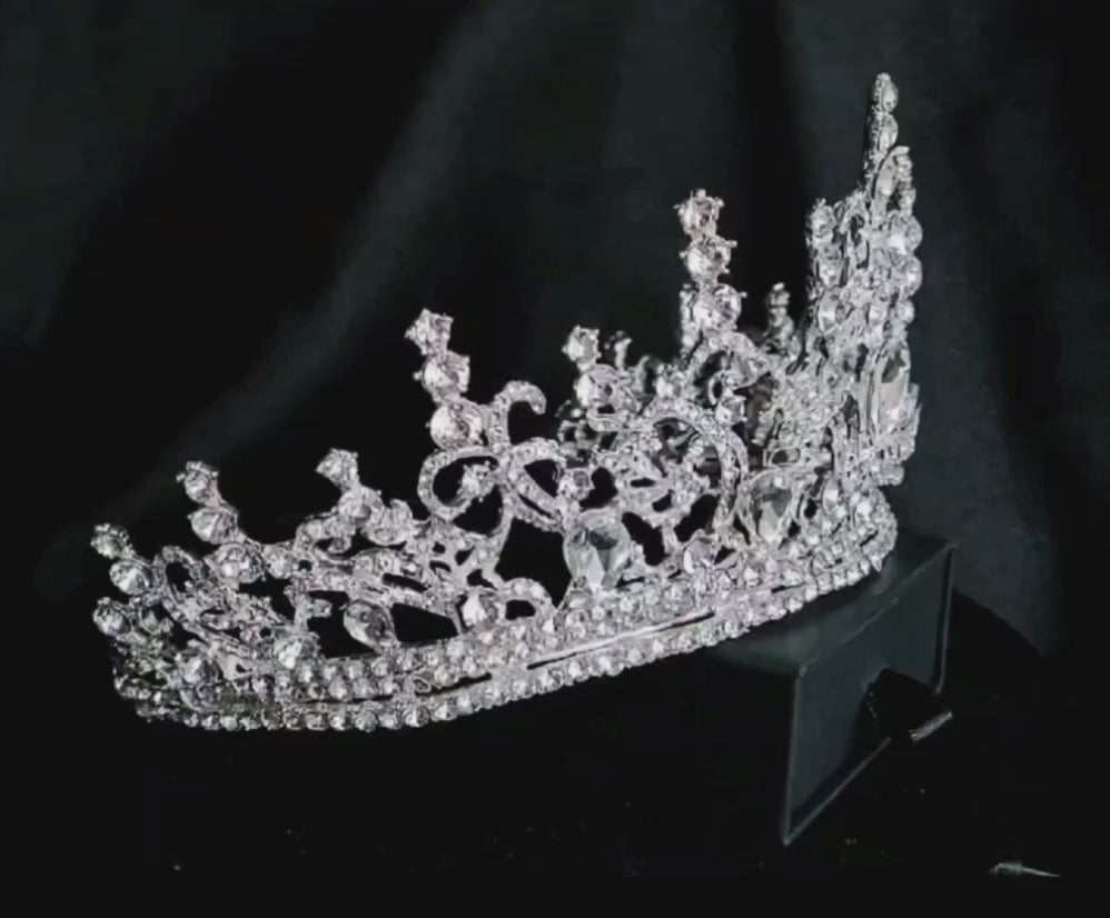 A close-up of a tiara with rhinestones on a black background. The tiara is made of silver and features a delicate design with cascading rhinestones. The rhinestones are clear and sparkling.