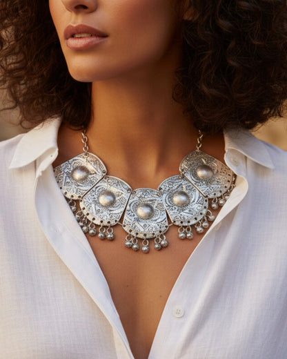 Detailed close-up of a large silver bohemian bib necklace featuring engraved circular medallions and hanging silver charms, styled with a white linen blouse.