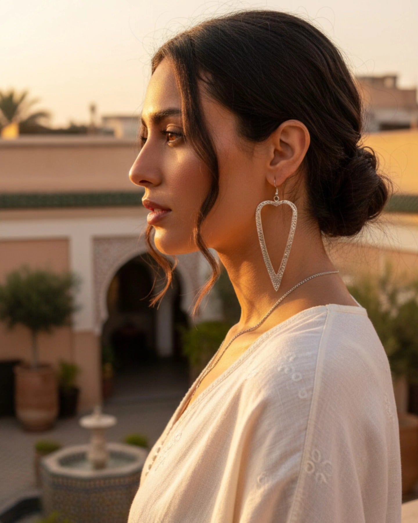 High-fashion side profile of a model wearing large, textured open-heart sterling silver earrings, featuring an elegant updo and sophisticated cream apparel in warm sunset lighting.