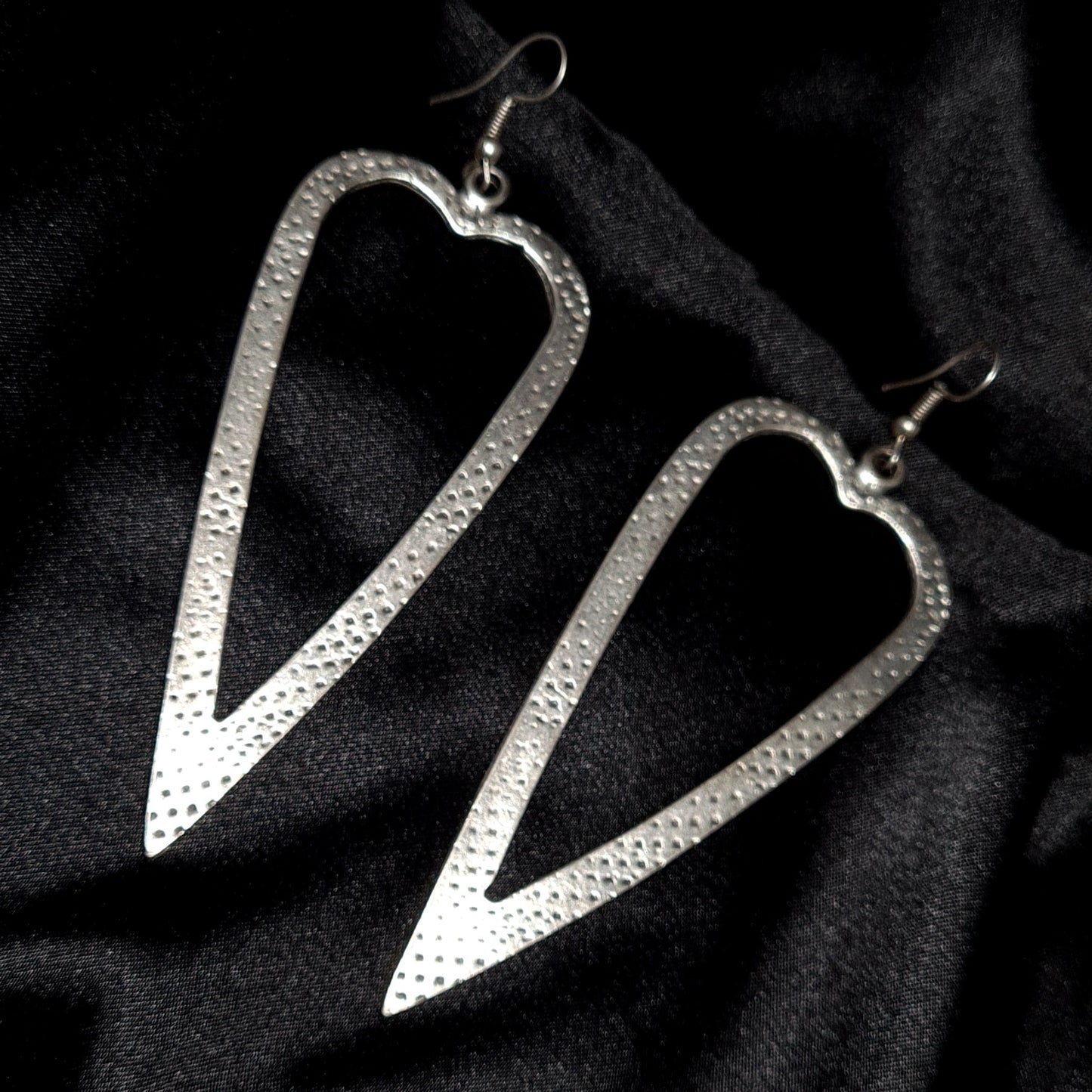Pair of oversized sterling silver open-heart earrings with a shimmering hammered texture, displayed on a deep black background with high-contrast luxury lighting.