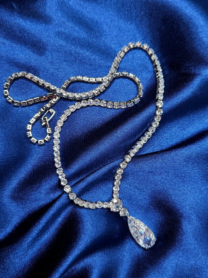 High-end luxury jewelry photography of the Tear Necklace resting on a rich, midnight blue fabric. The silver pendant and cubic zirconia stones shimmer against the deep textures, evoking a sense of premium elegance and sophisticated branding.