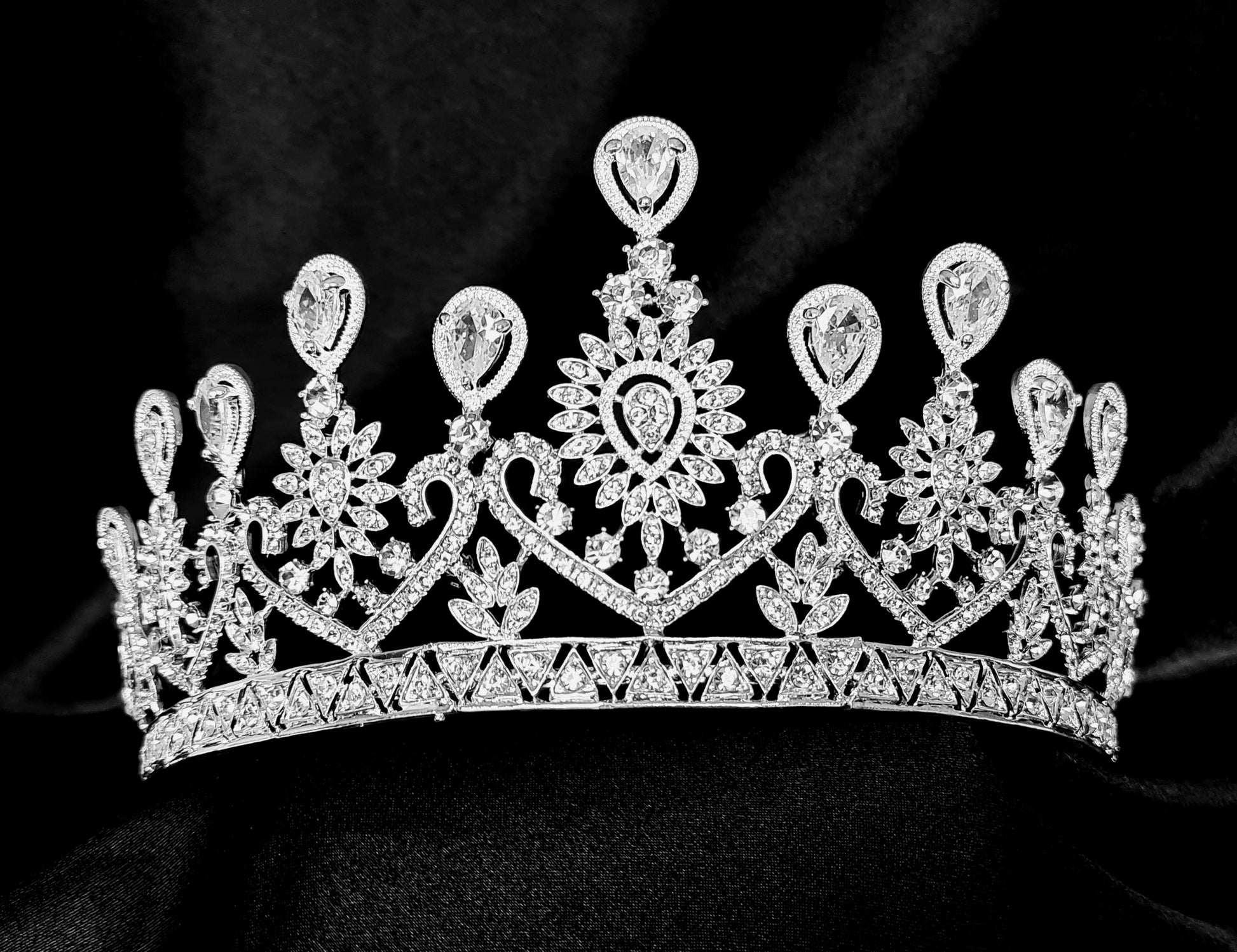 A close-up of a crown with diamonds on a black background. The crown is made of silver and is decorated with cubic zirconia stones, The stones are arranged in a pattern that resembles a flower, The crown is sitting on a black background.