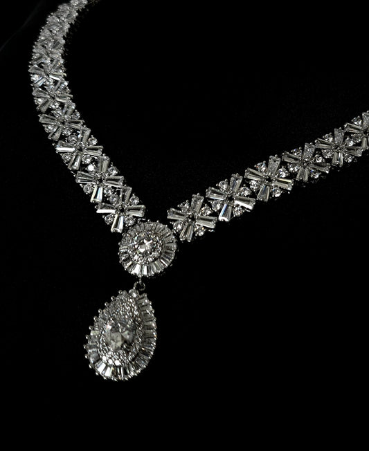 Image of a necklace with a large diamond pendant on a black background. The pendant is round and has a white diamond in the center. The necklace is made of silver and has a big chain. The background is black.