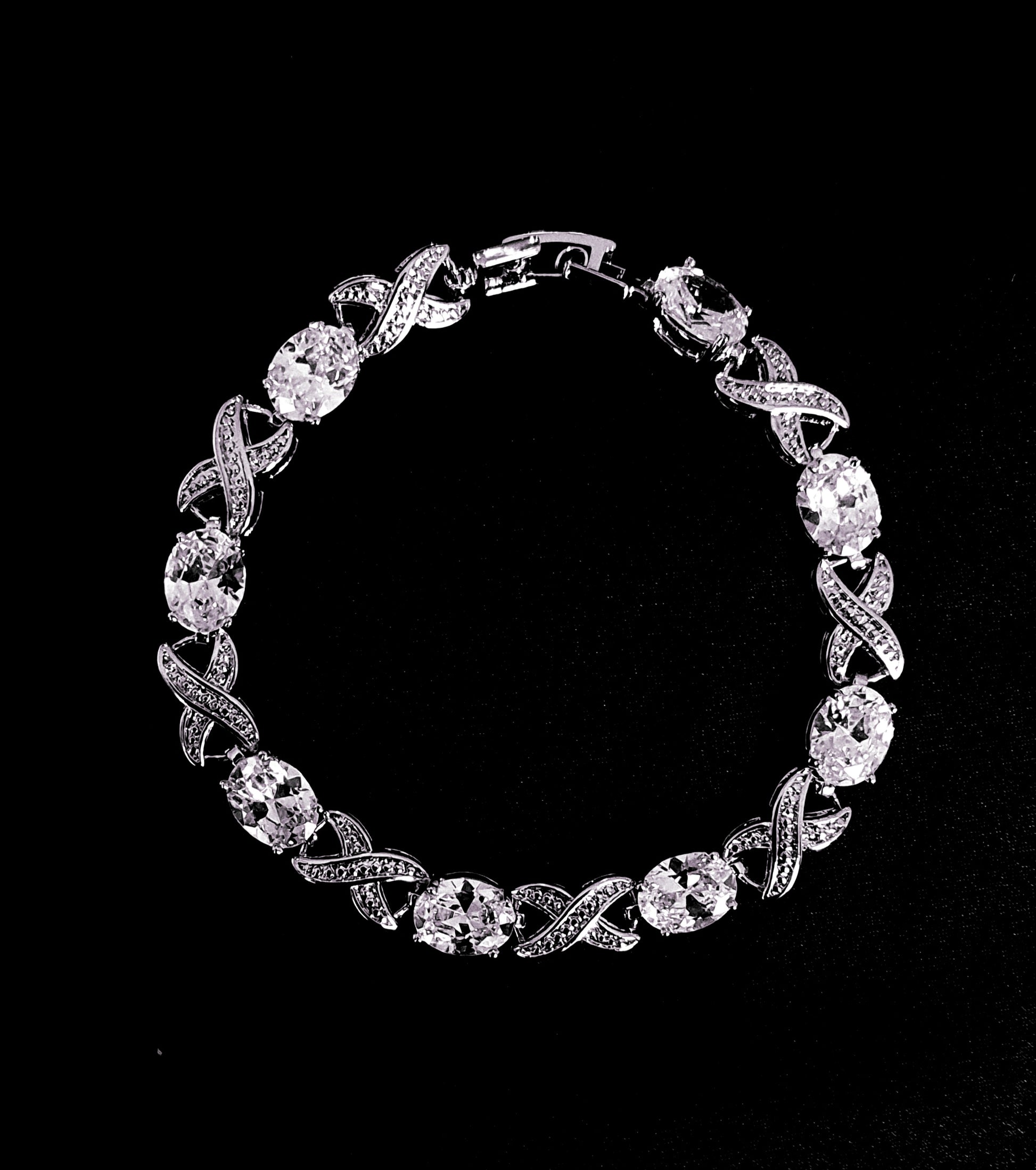 A close-up of a silver bracelet with a single row of white diamonds on a black background. The bracelet is simple in design and the diamonds are sparkling.