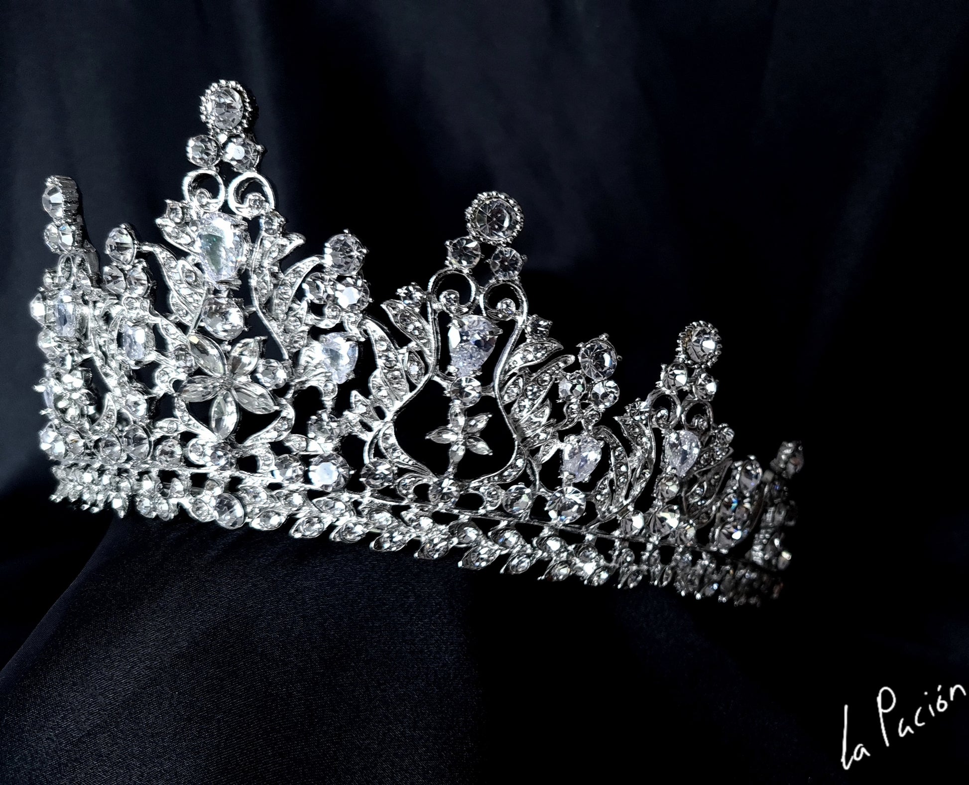 a tiara sitting on top of a black cloth. The tiara is silver in color and has a flower design. The rhinestones are clear and sparkling.