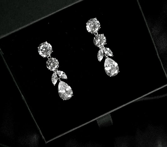 A pair of diamond earrings sitting on top of a black box. The earrings are teardrop-shaped and have a simple, elegant design. The diamonds are clear and sparkling