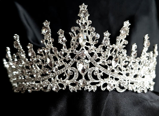 A close-up of a tiara with rhinestones on a black background. The tiara is made of silver and features a delicate design with cascading rhinestones. The rhinestones are clear and sparkling. 