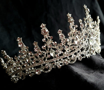 A close-up of a tiara with rhinestones on a black background. The tiara is made of silver and features a delicate design with cascading rhinestones. The rhinestones are clear and sparkling. 