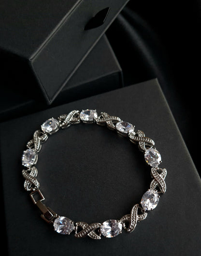 A close-up of a silver bracelet with a single row of white diamonds on a black background. The bracelet is simple in design and the diamonds are sparkling.