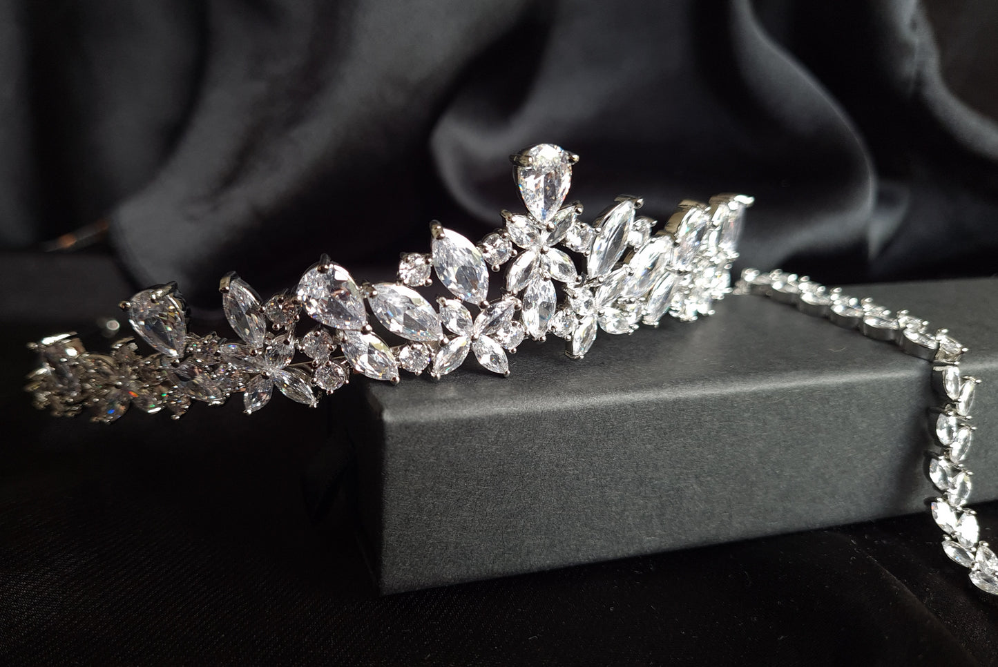 A tiara with diamonds on a black background. The tiara is made of silver and features a delicate design with cascading diamonds. The diamonds are clear and sparkling. The tiara is sitting on a table. next to a bracelet.