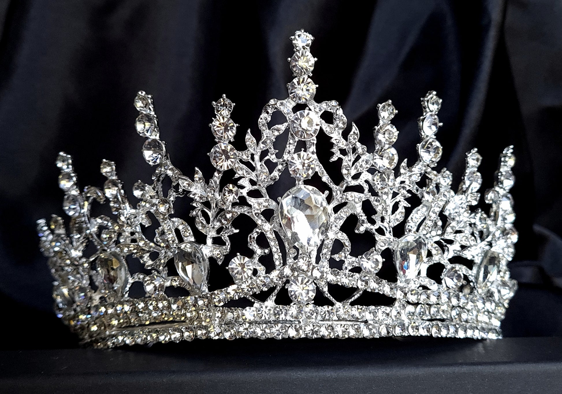  A close-up of a tiara with rhinestones on a black background. The tiara is made of silver and features a delicate design with cascading rhinestones. The rhinestones are clear and sparkling. 