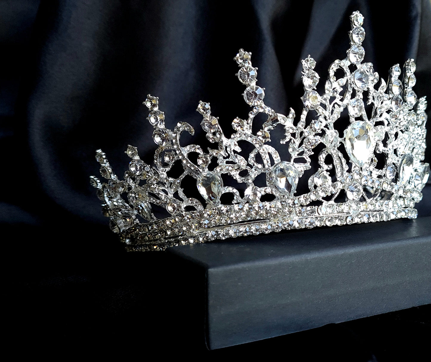  A close-up of a tiara with rhinestones on a black background. The tiara is made of silver and features a delicate design with cascading rhinestones. The rhinestones are clear and sparkling. 