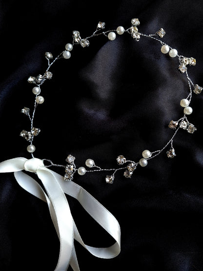 A wreath of pearls and crystals with a white ribbon. The wreath is 33 cm in diameter. It has a delicate design with pearls, crystals, and a white ribbon. The pearls are white and the crystals are clear. The background is black.