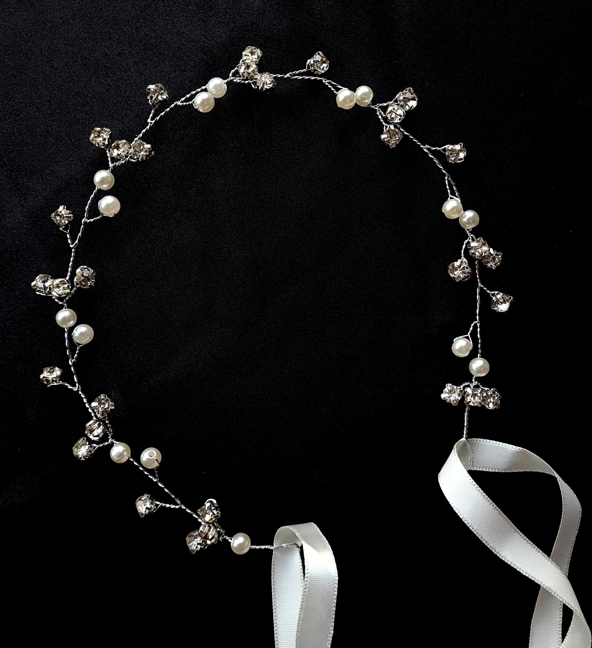 A wreath of pearls and crystals with a white ribbon. The wreath is 33 cm in diameter. It has a delicate design with pearls, crystals, and a white ribbon. The pearls are white and the crystals are clear. The background is black.