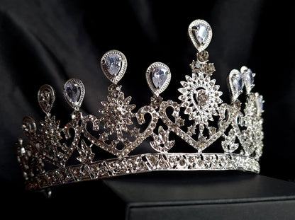 A close-up of a crown with diamonds on a black background. The crown is made of silver and is decorated with cubic zirconia stones, The stones are arranged in a pattern that resembles a flower,The crown is sitting on a black background.