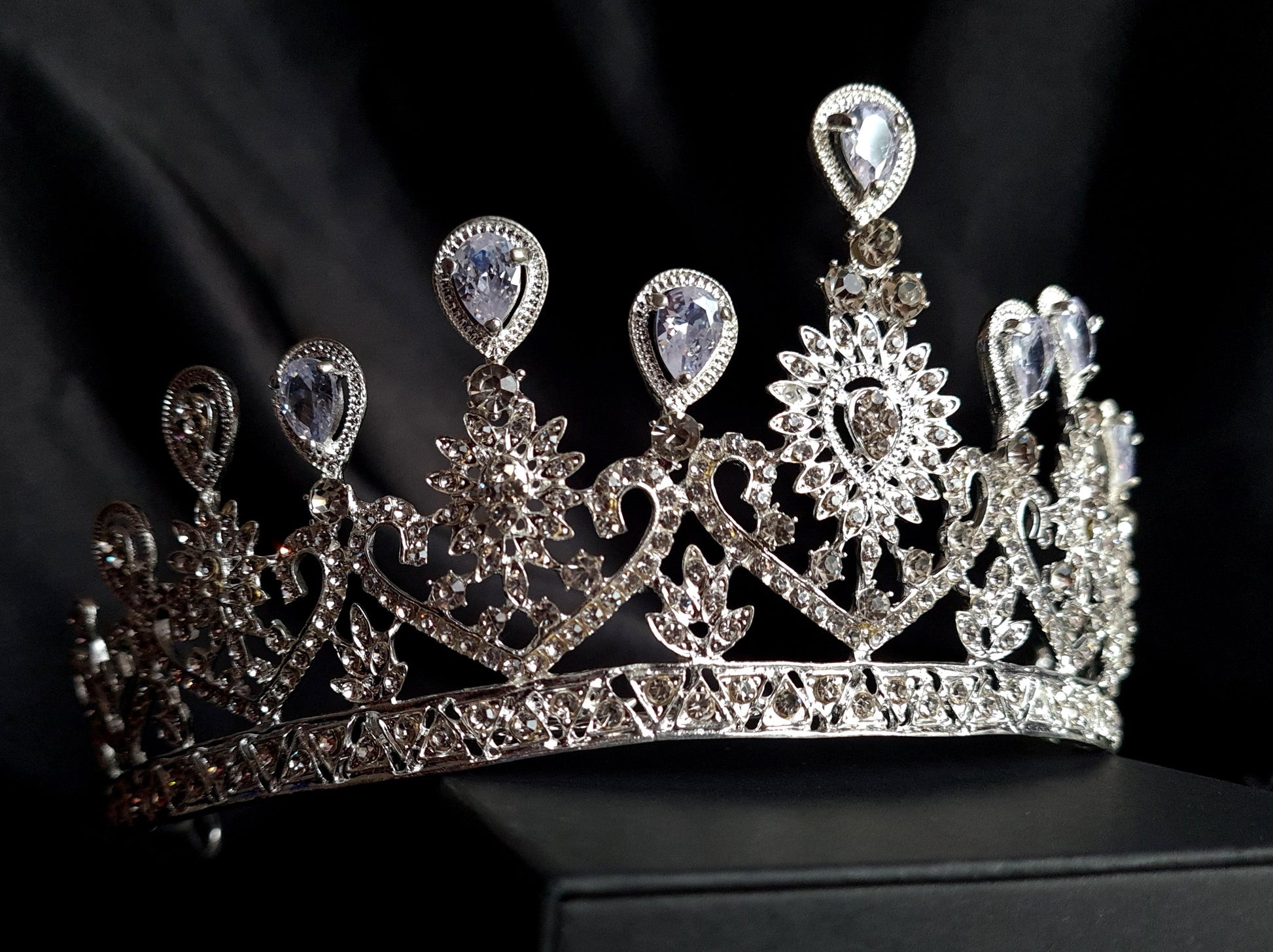 A close-up of a crown with diamonds on a black background. The crown is made of silver and is decorated with cubic zirconia stones, The stones are arranged in a pattern that resembles a flower,The crown is sitting on a black background.