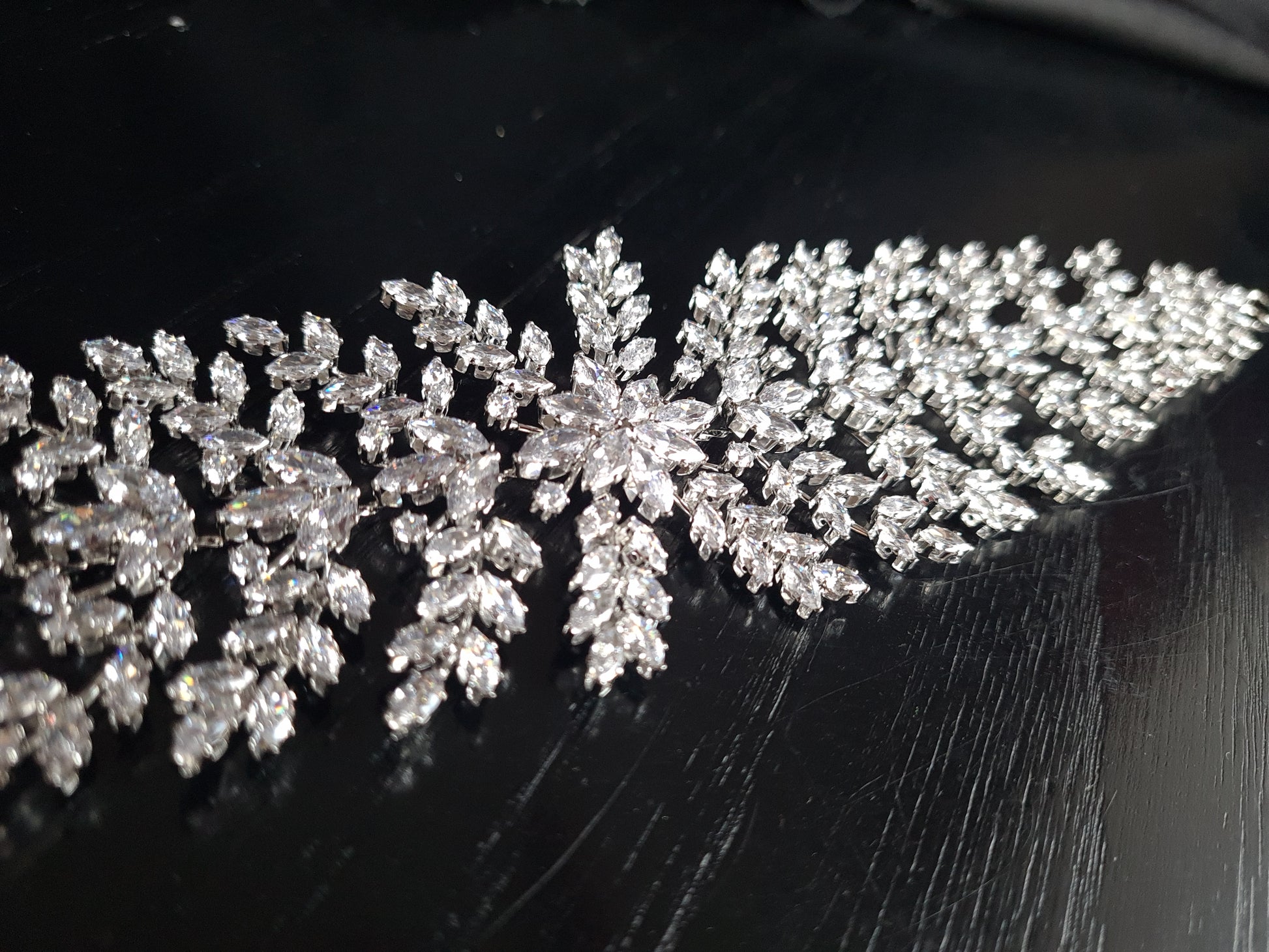 A wedding hair accessory made of metal and diamonds. The tiara has a delicate design with leaves and flowers. The diamonds are clear and sparkling. The background is black.