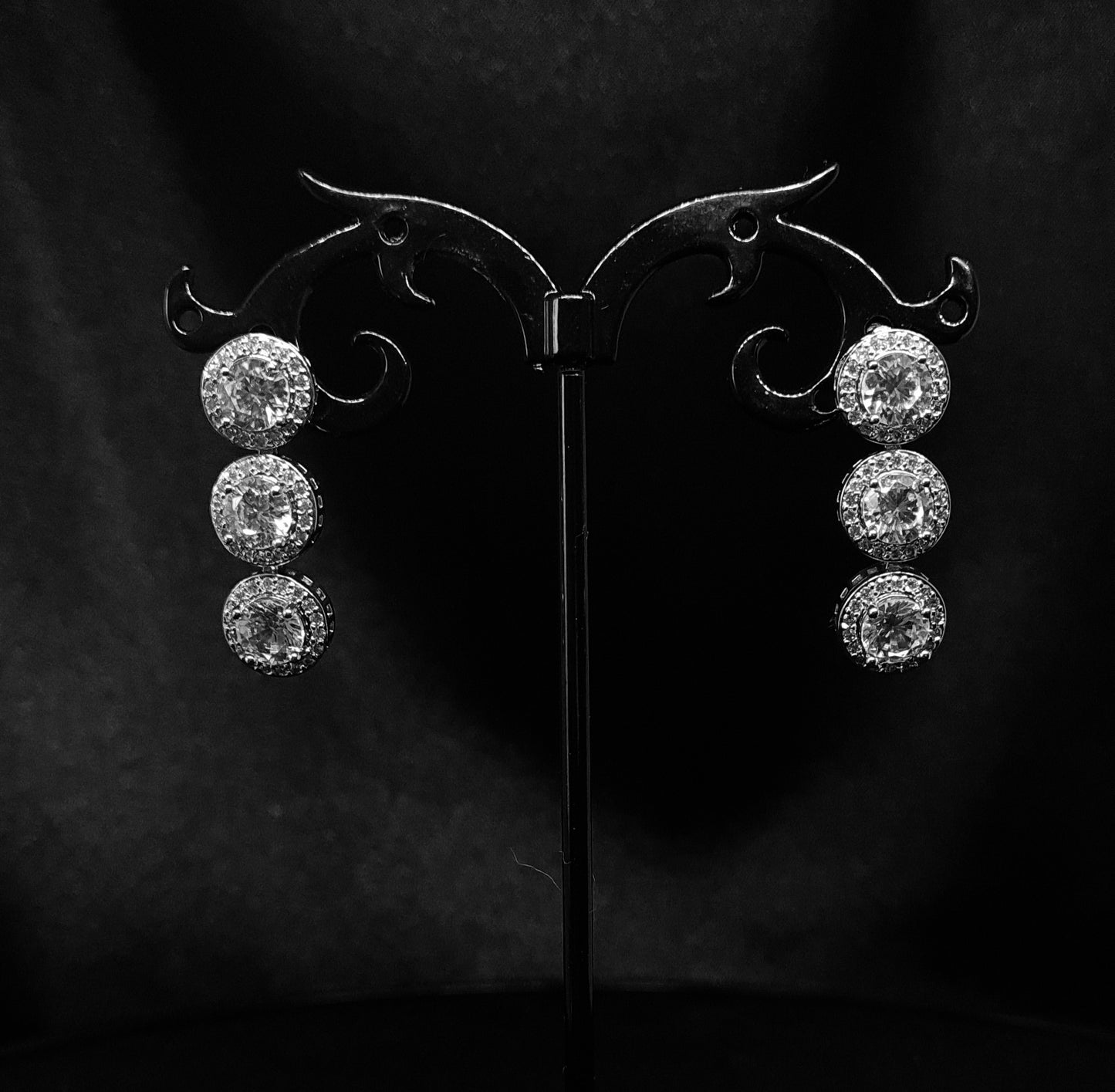 A pair of dangle earrings made of cubic zirconia. The earrings are round-shaped and have a simple, elegant design. The cubic zirconia stones are clear and sparkling. The earrings are hanging from a black metal wire.