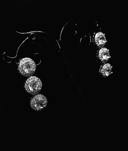 A pair of dangle earrings made of cubic zirconia. The earrings are round-shaped and have a simple, elegant design. The cubic zirconia stones are clear and sparkling. The earrings are hanging from a black metal wire.