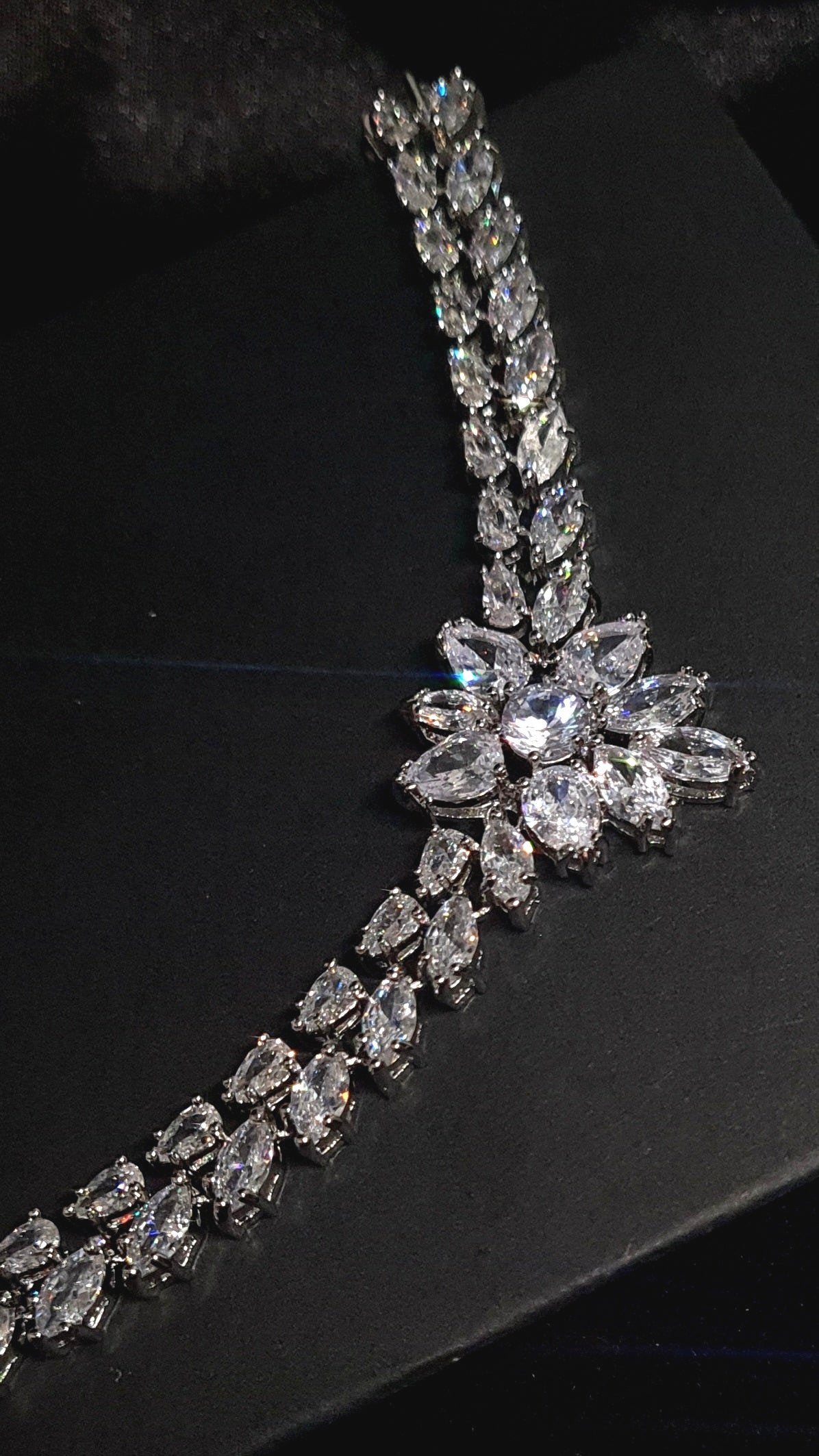 A close-up of a necklace on a black background. The necklace is made of silver and has diamonds in a variety of shapes and sizes. The diamonds are the main focus of the image and they sparkle in the light.