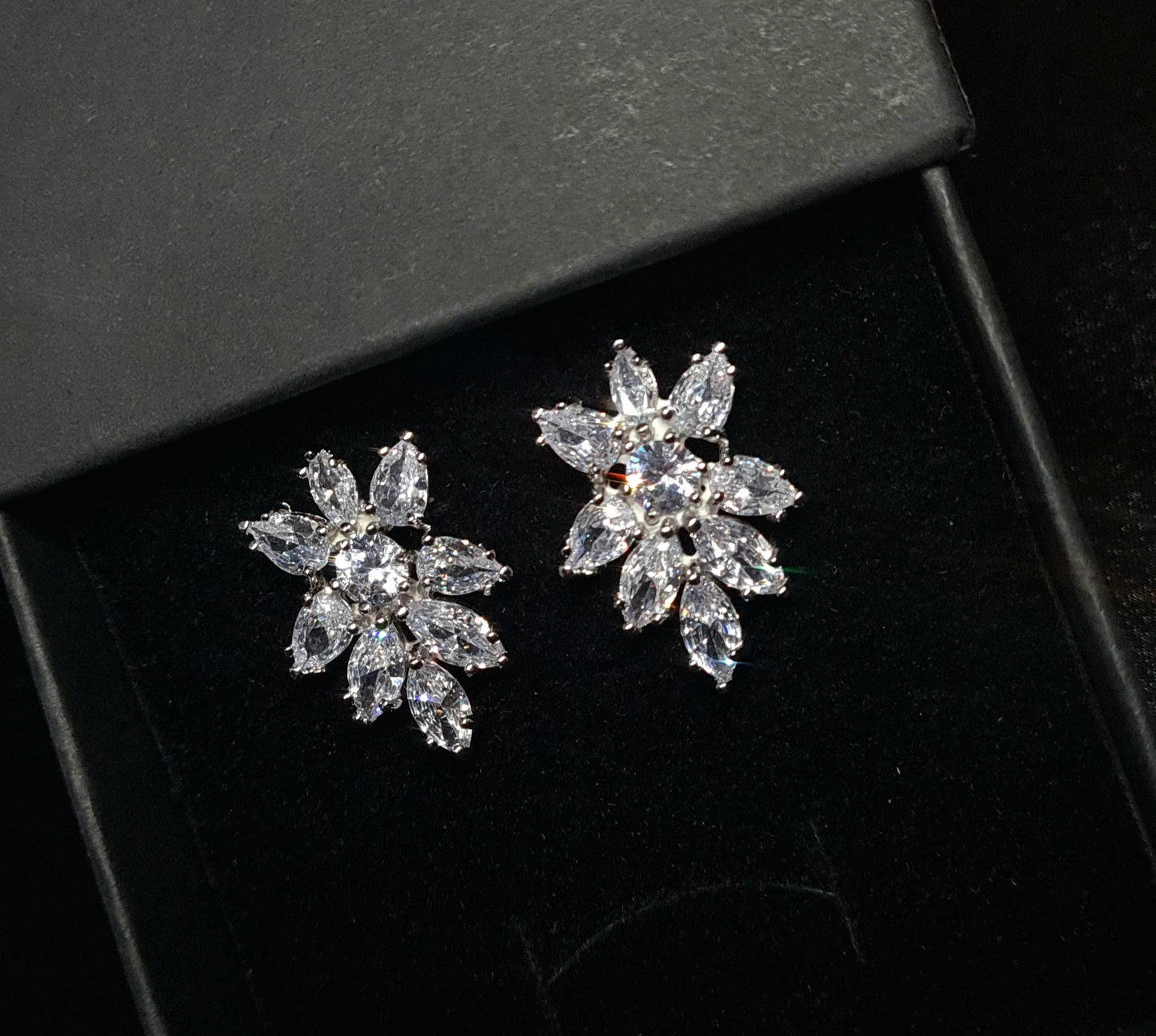 A pair of earrings in the shape of a flower, made from cubic zirconia stones. The earrings are sitting on a black surface. The flower is about 2.6 cm and is made up of petals that are clear and sparkling. The center of the flower is a large cubic zirconia stone.